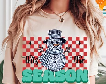 Może przedstawiać: Beżowa koszulka z motywem bałwana. Bałwanek wykonany jest z białych kropek i nosi cylinder i szalik. Napis "This The Season" jest wydrukowany na koszulce w kolorze czarnym i turkusowym.