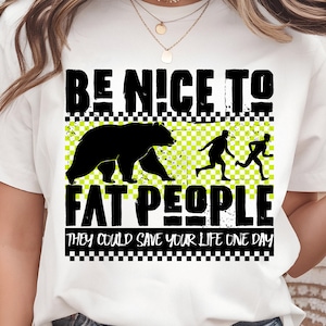Puede incluir: Camiseta blanca con un diseño gráfico negro. El diseño incluye el texto "BE NICE TO FAT PEOPLE" sobre una silueta de oso negro y dos figuras corriendo. Debajo, el texto "THEY COULD SAVE YOUR LIFE ONE DAY".