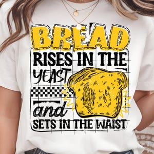 May include: White t-shirt with a yellow and black graphic. The graphic features the word "BREAD" in yellow, along with the phrases "RISES IN THE YEAST" and "and SETS IN THE WAIST". A yellow illustration of a slice of bread is also included.