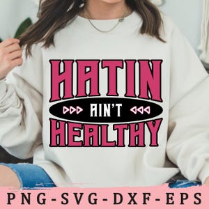 May include: A white sweatshirt with pink text that reads "Hatin' Ain't Healthy" in a black oval with pink triangles on either side.