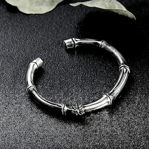 Puede incluir: Pulsera plateada estilo bambú con un pequeño acento dorado. La pulsera es abierta y descansa sobre una superficie oscura y texturizada. El diseño imita la estructura segmentada de los tallos de bambú, creando una estética naturalista.