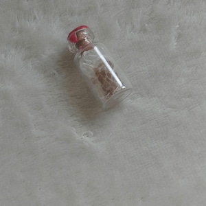 Op de afbeelding: Een kleine, heldere glazen fles met een rode stopper. De fles bevat een lichtbruine substantie. De fles ligt op een witte, zachte ondergrond.