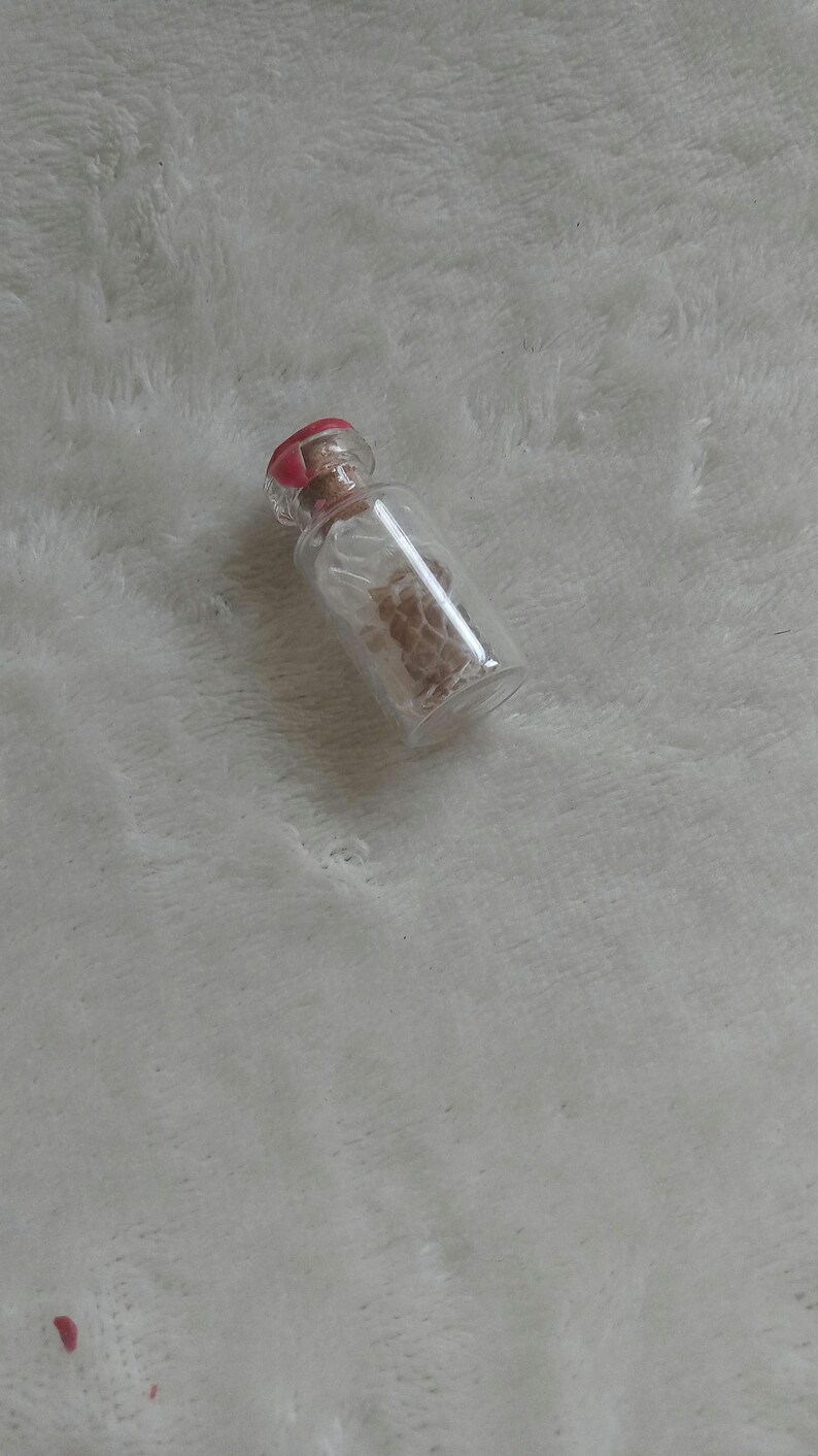 Op de afbeelding: Een kleine, heldere glazen fles met een rode stopper. De fles bevat een lichtbruine substantie. De fles ligt op een witte, zachte ondergrond.