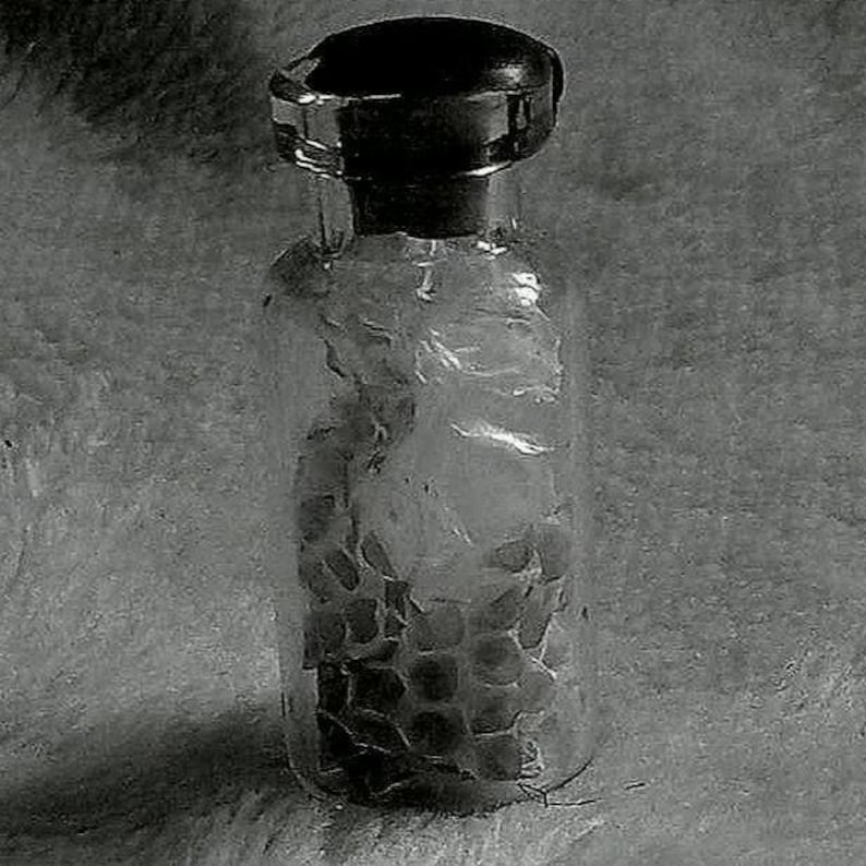 Op de afbeelding: Een heldere glazen fles met een donkere, afgeronde stopper. De fles bevat een textuurrijke, donkergekleurde substantie. De fles is cilindrisch met een licht getextureerd oppervlak. De stopper heeft een decoratief, geribbeld ontwerp.