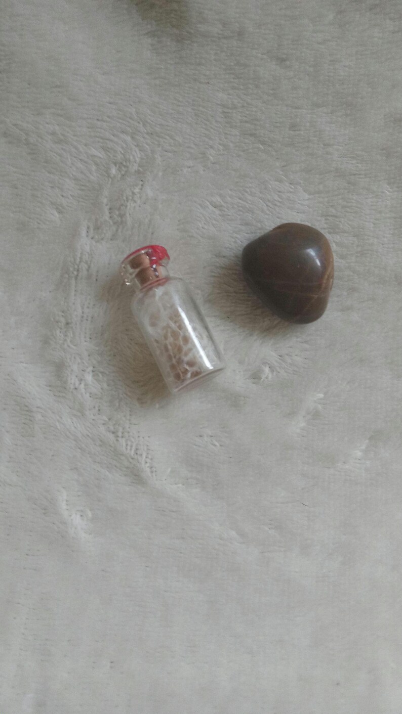 Op de afbeelding: Een kleine, heldere glazen fles met een rood gekleurde kurk, gevuld met een lichtgekleurde substantie, en een gladde, bruine, hartvormige steen. De items zijn geplaatst tegen een zachte, witte, getextureerde achtergrond.