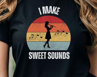 Może przedstawiać: Czarny T-shirt z grafiką w stylu retro przedstawiającą kobietę grającą na flecie. Grafika zawiera koło w kolorze zachodzącego słońca z nutami muzycznymi i tekstem "I MAKE SWEET SOUNDS".
