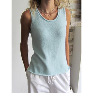 May include: A light blue sleeveless knit tank top with a round neckline and a simple, straight hem. The garment features a fine, uniform knit texture and is worn with white drawstring pants and layered metallic necklaces around the neck.