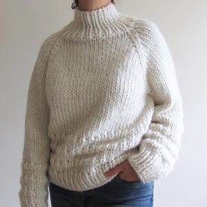 Puede incluir: Un suéter de punto blanco con cuello alto y mangas largas. El suéter tiene un corte holgado y una longitud ligeramente recortada.