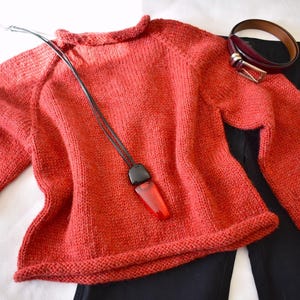 Puede incluir: Un su&eacute;ter de punto color coral con mangas largas, un collar negro con un colgante rojo, un cintur&oacute;n burdeos y pantalones negros se muestran. El su&eacute;ter es el punto focal, con los otros art&iacute;culos dispuestos alrededor.