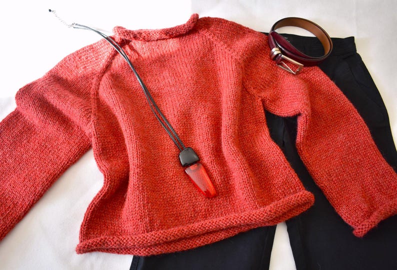 Puede incluir: Un su&eacute;ter de punto color coral con mangas largas, un collar negro con un colgante rojo, un cintur&oacute;n burdeos y pantalones negros se muestran. El su&eacute;ter es el punto focal, con los otros art&iacute;culos dispuestos alrededor.