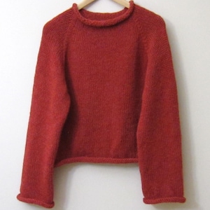 Puede incluir: Un jersey de punto de color rojo ladrillo con cuello redondo y mangas largas. El jersey est&aacute; hecho de un hilo texturizado y tiene pu&ntilde;os y dobladillo acanalados. La prenda se exhibe en una percha de madera sobre un fondo blanco.