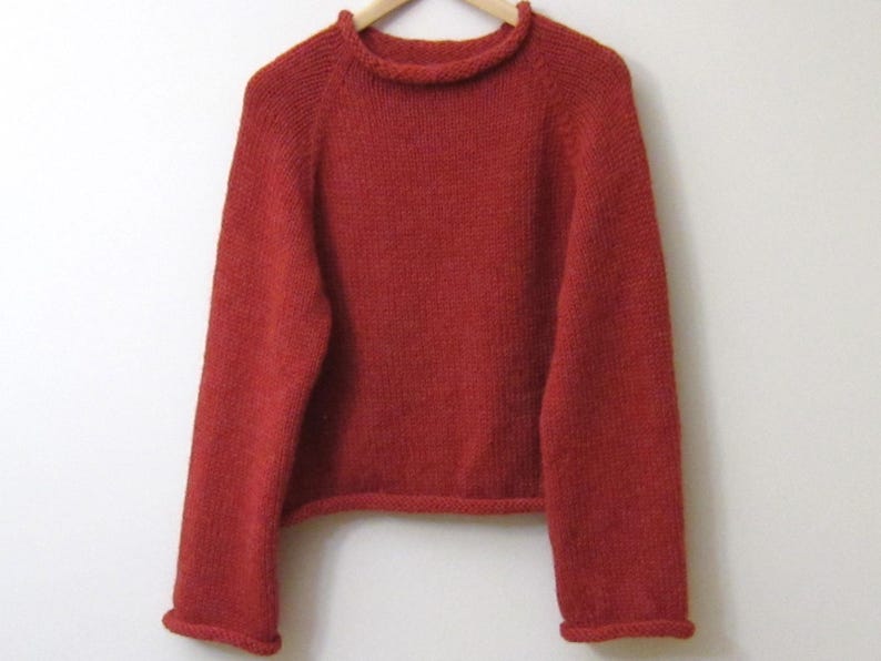 Puede incluir: Un jersey de punto de color rojo ladrillo con cuello redondo y mangas largas. El jersey est&aacute; hecho de un hilo texturizado y tiene pu&ntilde;os y dobladillo acanalados. La prenda se exhibe en una percha de madera sobre un fondo blanco.