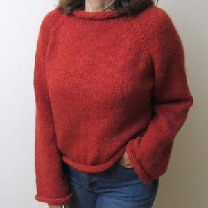 May include: A red knit sweater with a round neckline and long sleeves. The sweater has a ribbed hem and cuffs. The person is wearing blue jeans. The sweater is a solid color and appears to be handmade.