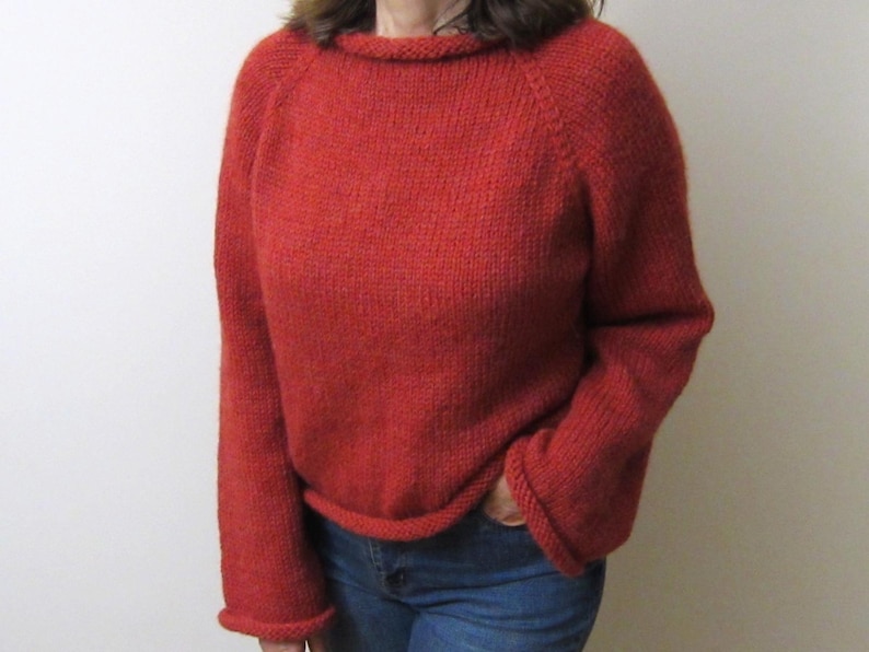 Puede incluir: Un su&eacute;ter rojo tejido con cuello redondo y mangas largas. El su&eacute;ter tiene dobladillo y pu&ntilde;os acanalados. La persona lleva vaqueros azules. El su&eacute;ter es de color liso y parece hecho a mano.