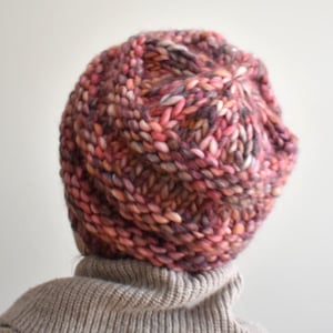 May include: A hand-knitted beanie in a mix of pink, purple, and peach hues. The chunky knit hat is made of thick yarn and has a ribbed brim. The hat is shown from the back, with a neutral-coloured turtleneck sweater visible.