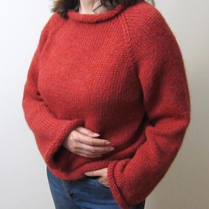 Puede incluir: Un su&eacute;ter rojo tejido con cuello redondo y mangas largas. El su&eacute;ter es de color liso y parece hecho a mano. Las mangas tienen un dise&ntilde;o acampanado en los pu&ntilde;os. La persona lleva vaqueros azules.