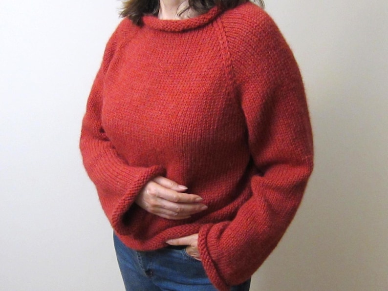 Puede incluir: Un su&eacute;ter rojo tejido con cuello redondo y mangas largas. El su&eacute;ter es de color liso y parece hecho a mano. Las mangas tienen un dise&ntilde;o acampanado en los pu&ntilde;os. La persona lleva vaqueros azules.