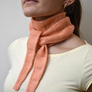 May include: A peach-coloured knitted scarf with a textured pattern. The scarf is tied around the neck, with the ends hanging down. The scarf is made of a soft, warm material, ideal for cooler weather.