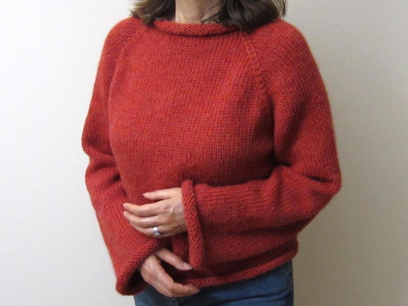 Puede incluir: Un su&eacute;ter de punto color &oacute;xido con cuello redondo y mangas largas. Las mangas tienen un dise&ntilde;o acampanado en los pu&ntilde;os. El su&eacute;ter est&aacute; hecho de un hilo grueso y texturizado y parece hecho a mano. El modelo lleva vaqueros azules.