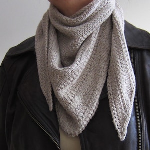 May include: A light beige knitted scarf is worn around the neck. The scarf has a triangular shape with a textured pattern and a decorative edge. The person is wearing a dark brown leather jacket.
