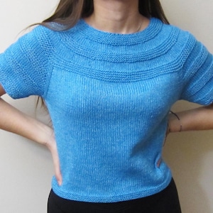 Könnte beinhalten: Ein hellblaues Strickoberteil mit kurzen Ärmeln und einem runden Ausschnitt. Das Oberteil hat ein strukturiertes Strickmuster und eine leicht verkürzte Länge.