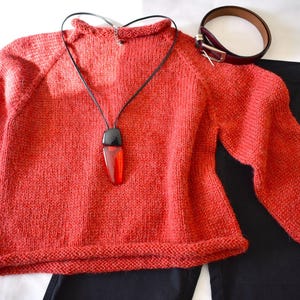 Puede incluir: Un su&eacute;ter de punto color coral con mangas largas, combinado con pantalones negros y un cintur&oacute;n marr&oacute;n. Un collar negro con un colgante rojo y negro est&aacute; drapeado sobre el su&eacute;ter. El conjunto est&aacute; dispuesto sobre una superficie blanca.
