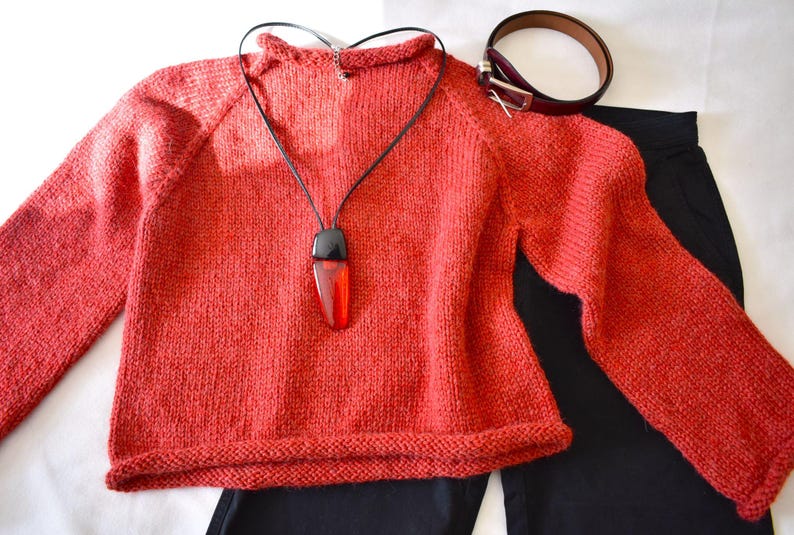 Puede incluir: Un su&eacute;ter de punto color coral con mangas largas, combinado con pantalones negros y un cintur&oacute;n marr&oacute;n. Un collar negro con un colgante rojo y negro est&aacute; drapeado sobre el su&eacute;ter. El conjunto est&aacute; dispuesto sobre una superficie blanca.