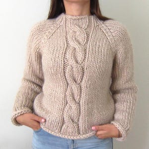 Può includere: Un maglione di lana beige con un motivo a treccia. Il maglione ha un collo rotondo e maniche lunghe.