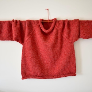 Puede incluir: Un jersey rojo tejido con un cuello ancho y mangas largas. El jersey se muestra en una barra de madera sobre un fondo blanco. Las mangas miden aproximadamente 61 cm de largo.