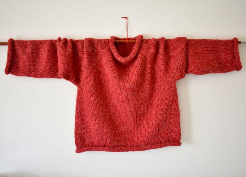 Puede incluir: Un jersey rojo tejido con un cuello ancho y mangas largas. El jersey se muestra en una barra de madera sobre un fondo blanco. Las mangas miden aproximadamente 61 cm de largo.