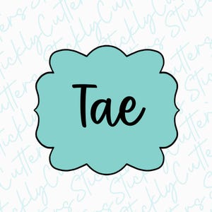 May include: A light blue cookie cutter with a scalloped edge and the name "Tae" written in black cursive.