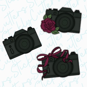 May include: Three black camera cookie cutters with different designs. One camera has a red rose with green leaves, another has a red bow, and the third is plain.