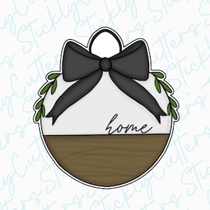 May include: A round, white ornament with a black bow and green leaves. The ornament has the word "home" written on it and a brown wooden base.