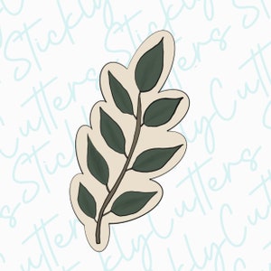 May include: A green leaf cookie cutter with a beige stem and multiple leaves. The cookie cutter is isolated on a white background.