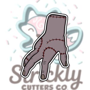 May include: A gray hand cookie cutter with red stitching details. The hand has a white outline and is set against a blurred background with the text "Strickly Cutters Co."