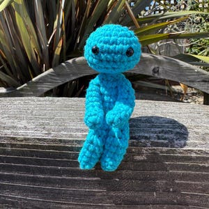 May include: A turquoise blue crochet doll with a round head and small black eyes. The doll has a simple, humanoid form with arms and legs, and is posed standing on a wooden surface. The doll is a whimsical, soft toy, likely handmade.