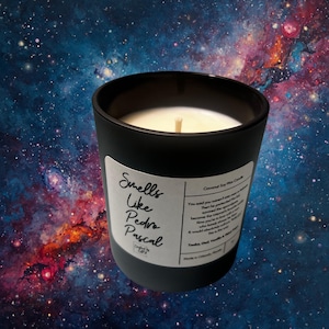 May include: A black candle in a glass jar with a white label that reads "Smells Like Pedro Pascal." The candle has a white wax and a wooden wick. The background is a colorful galaxy.