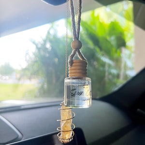 May include: A car air freshener with a clear glass bottle and a wooden cap. The bottle is hanging from a rearview mirror with a gold chain and a brown rope. The bottle is labeled "Spun Cotton" and has a clear liquid inside. A gold crystal is hanging from the chain below the bottle.