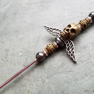 May include: A silver and gold metal skewer with a skull and wings design. The skewer has black beads and a silver metal tip.