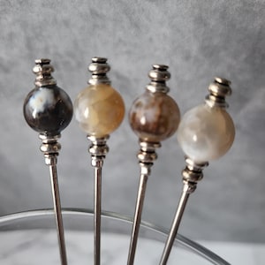May include: Four silver cocktail picks with agate stone tops. The stones are in shades of brown, white, and gray.