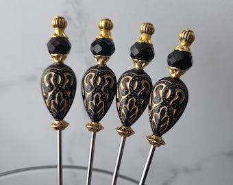 Puede incluir: Cuatro palillos de cóctel decorativos. Cada palillo presenta una cuenta negra en forma de lágrima con filigrana dorada, rematada con una cuenta facetada negra y una terminación dorada. Los palillos tienen largos y delgados tallos plateados.