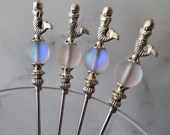 Könnte beinhalten: Vier silberne Cocktailspieße mit Meerjungfrau-Aufsätzen. Jeder Spieß hat eine silberne Meerjungfrau-Figur auf einer mattierten Glaskugel mit schillernden Farbtönen. Die Spieße sind fächerförmig angeordnet, was auf ihre Verwendung hindeutet.