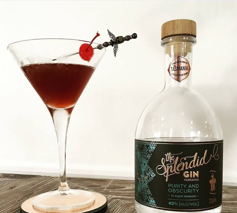May include: A clear glass bottle of The Splendid Gin from Tasmania. The bottle has a black label with gold lettering and a teal geometric pattern. The label reads "The Splendid Gin, Tasmania, Purity and Obscurity - in equal measure - 42% [ALC/VOL] 700ml".
