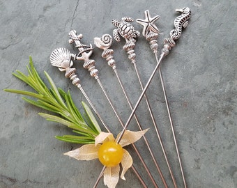 Könnte beinhalten: Sechs silberne Cocktailspieße mit maritimen Motiven: Muschel, Anker, Schnecke, Schildkröte, Seestern und Seepferdchen. Auf einer grauen Oberfläche mit Rosmarinzweig und gelber Frucht angeordnet.