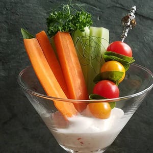 May include: A clear glass martini glass filled with a white dip and topped with fresh vegetables. The vegetables include celery sticks, carrot sticks, and cherry tomatoes. A skewer with a decorative bead holds a skewer of cherry tomatoes and yellow grape tomatoes.