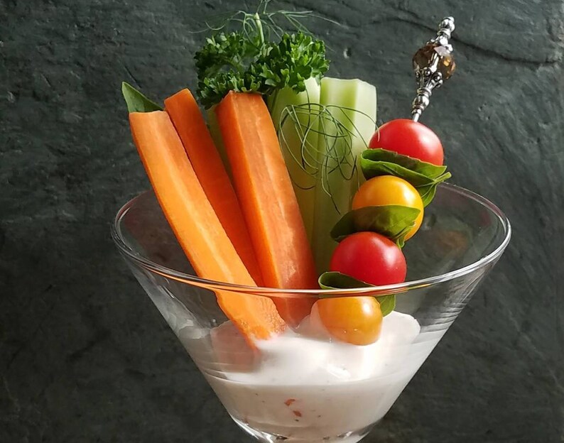May include: A clear glass martini glass filled with a white dip and topped with fresh vegetables. The vegetables include celery sticks, carrot sticks, and cherry tomatoes. A skewer with a decorative bead holds a skewer of cherry tomatoes and yellow grape tomatoes.