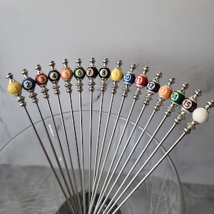 May include: Fifteen cocktail picks with silver stems and numbered billiard ball tops. The balls are in various colours, including yellow, blue, red, and green. The picks are arranged in a fan shape, suggesting they are ready for use.