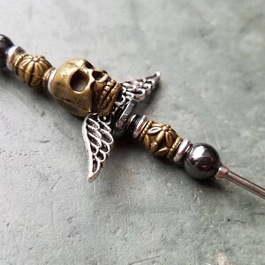 May include: A silver and gold metal skull with wings attached to a long, thin metal rod with black beads.