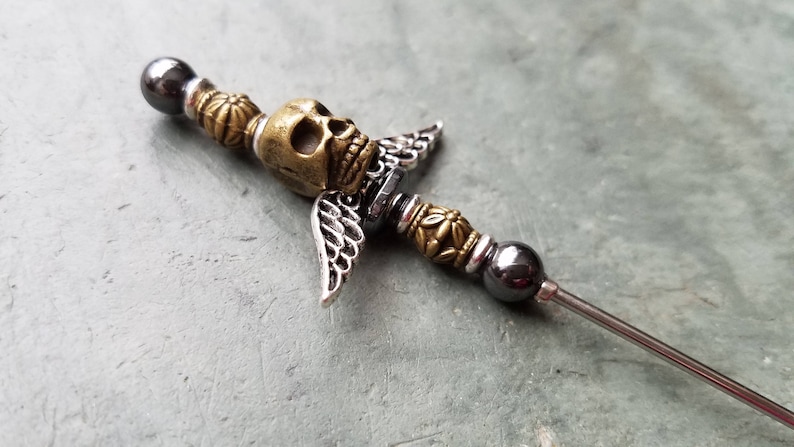 May include: A silver and gold metal skull with wings attached to a long, thin metal rod with black beads.