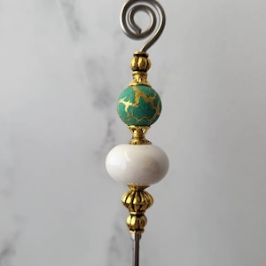 May include: A decorative metal stick with a gold-colored top and a white and green bead. The top of the stick has a spiral design.